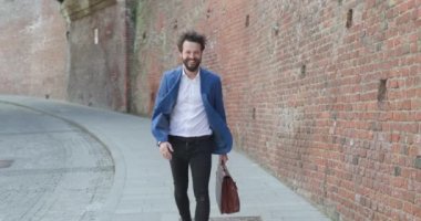 Uzun bıyıklı ve sakallı çekici bir genç. Elinde bavul, kameraya doğru yürüyor ve dışarıda gülümsüyor.