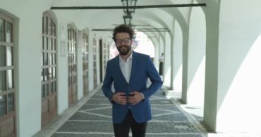 Sakallı, uzun bıyıklı ve gözlüklü genç bir adam ceket düğmesine el ele tutuşuyor ve şehrin arka planında yürüyor, yürüyor ve gülümsüyor.