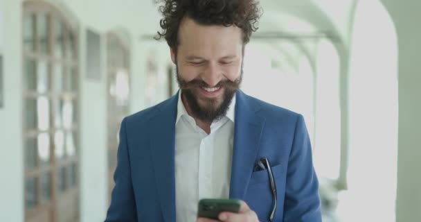 微笑的卷发男人拿着咖啡 低头看电话 看邮件和短信 当相机靠近外面的时候笑 — 图库视频影像