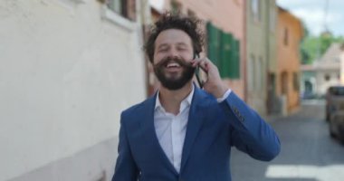 Çekici kıvırcık saçlı adam yürüyor, telefona sarılıyor, harika bir konuşma yapıyor ve dışarıda coşkuyla konuşuyor.