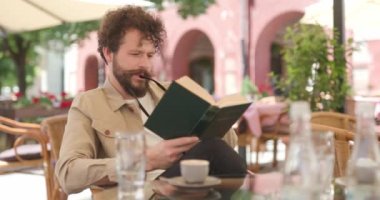 Gözlüklerini dudaklarına dayayıp kitap okuyan ve kameraya gülümseyen çekici, sıradan bir adam.