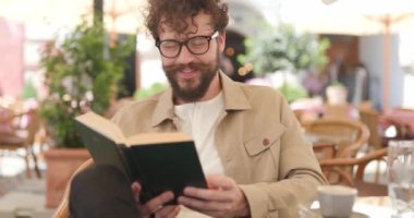 Genç adam kitabını okuyor, kameraya bakıyor, gülümsüyor, gözlük takıyor ve terasta oturuyor.