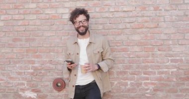 Çekici, sıradan bir adam, tuğla bir duvara yaslanmış, gözlük takmış, elinde telefon ve bir fincan kahveyle eğleniyor.