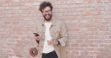 Tuğla duvara yaslanmış sıradan bir adam, kahve fincanını kaldırıyor ve içiyor, eline telefon alıyor ve gözlük takıyor.