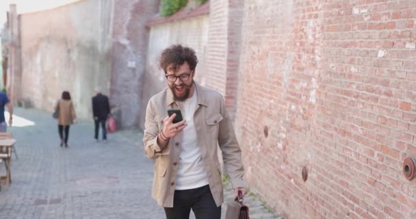携帯電話とブリーフケースを持ってシビウの旧市街を歩くハンサムなカジュアルな男は ジャンプして成功を祝う — ストック動画