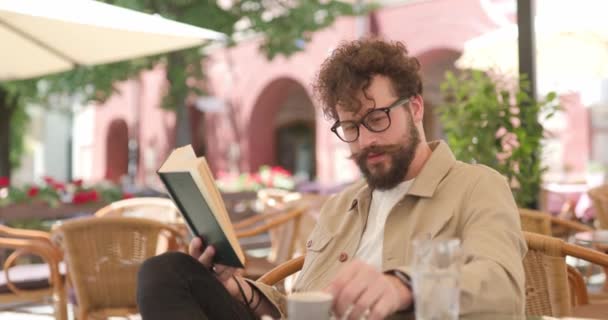 Młodzieniec Czytający Książkę Pijący Kawę Noszący Okulary Siedzący Tarasie — Wideo stockowe