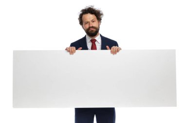 Çekici iş adamı elinde boş bir ilan panosu tutuyor ve beyaz arka planda huysuz hissediyor.