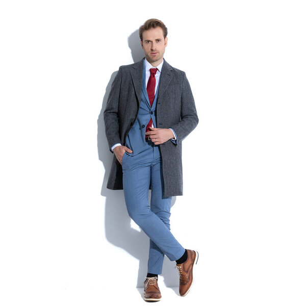 крутой бизнесмен скрестил ноги, держа одну руку в кармане и прикрывая пальто на белом фоне