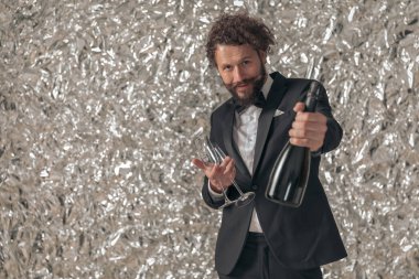 Zarif smokinli yakışıklı adam şampanya şişesi tavsiye ediyor, bardak tutuyor ve stüdyoda folyo arka planda kutlama yapıyor.