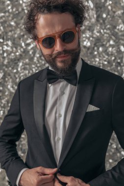 Siyah smokinli havalı damat ceketini ayarlıyor ve stüdyoda folyo arka planda poz verirken yan tarafa bakıyor.