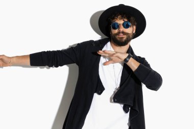 Genç adam ellerini yana kaydırıyor ve beyaz arka planda havalı bir şapka ve güneş gözlüğü takıyor.