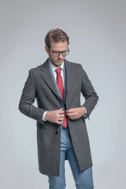 Dramatik iş adamı ceketini kapatıyor, aşağı bakıyor ve gri arka planda gözlük takıyor.
