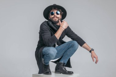 Havalı, çömelmiş, sıradan bir adam yüzüne dokunuyor ve gri arka plana karşı sert bir tutum sergiliyor.