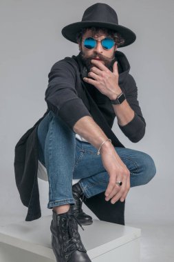 Sert tavırlı seksi bir adam yüzüne dokunuyor ve gri arka plana çömeliyor.