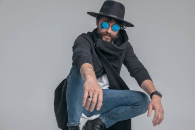 Güneş gözlüklü, şapkalı, çömelmiş ve gri arka planda poz veren seksi bir adam.