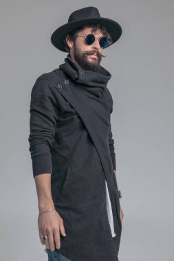 Gri stüdyo arka planında hırka, güneş gözlüğü ve şapka takan çekici bir adam.