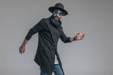 Seksi manken poz veriyor, güneş gözlüğü takıyor ve gri arka planda bir şapka takıyor.