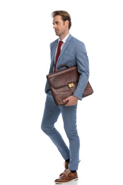 Mavi takım elbiseli havalı genç işadamının yan görünüşü. Stüdyoda beyaz arka planda tek başına yürürken bavulu tutmak ve yan bakmak.