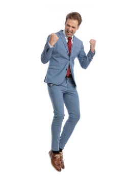 Heyecanlı, mavi takım elbiseli, yumruklarını havada tutan, zaferi kutlayan, havaya zıplayan, stüdyoda beyaz arka planda gülen ve bağıran zarif bir adam.