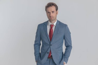 Mavi takım elbiseli yakışıklı, tıraşsız iş adamı. Stüdyoda gri arka planda elleri ceplerinde poz veriyor.