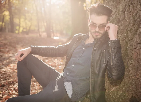 Jovem homem casual apoiando-se em uma árvore, fixando seus óculos . — Fotografia de Stock