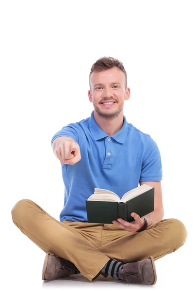 Young seated man with book points at you — Stock Photo, Image