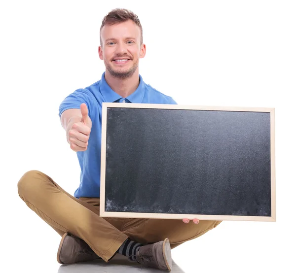 Young seated man with blackboard shows thumb up — Stock Photo, Image