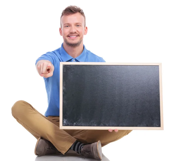 Young seated man with blackboard points at you — Stock Photo, Image