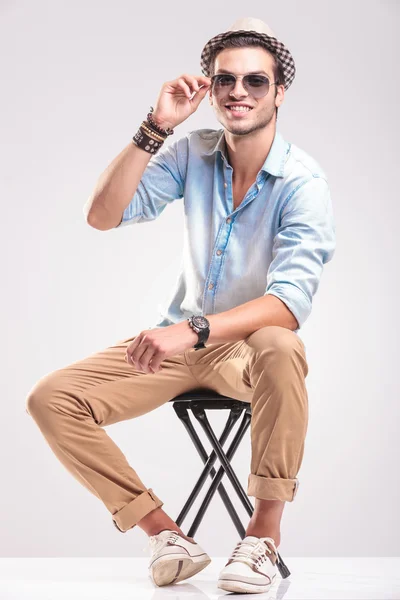 Sonriente hombre de la moda arreglando sus gafas de sol mientras está sentado — Foto de Stock