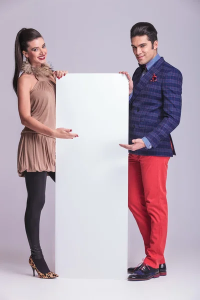 Fashion couple presenting a white empty bilboard. — Stock Photo, Image