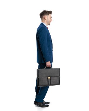 Lacivert takım elbiseli yakışıklı bir genç beyaz arka planda bavulu tutuyor ve yana bakıyor.