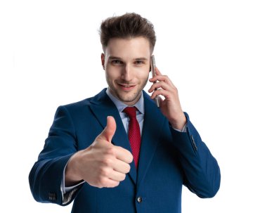 Lacivert takım elbiseli mutlu genç işadamı telefonda konuşuyor ve beyaz arka planda gülümserken bir jest gibi başparmak kaldırıyor.