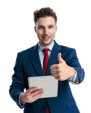 Lacivert takım elbiseli mutlu bir iş adamı beyaz arkaplan önünde gülümserken parmak hareketi yapıyor ve tableti tutuyor.