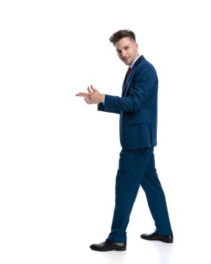 Lacivert takımlı, mutlu, zarif bir adamın yan görünüşü. Parmakla işaret ediyor ve beyaz arka planda gülümsüyor.