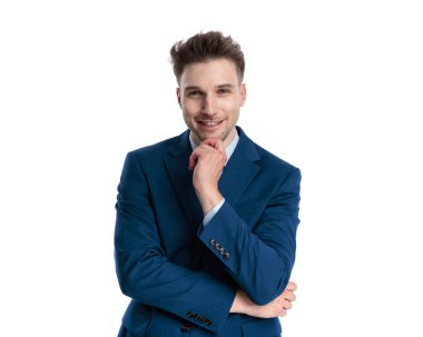 Lacivert takımlı seksi genç adam beyaz arka planda gülümserken çenesine dokunuyor ve düşünüyor.