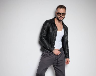 Siyah deri ceketli kibirli seksi bir adam. Güneş gözlüklü. Gri arka planda pantolonu tutarken kendine güvenen bir poz veriyor.