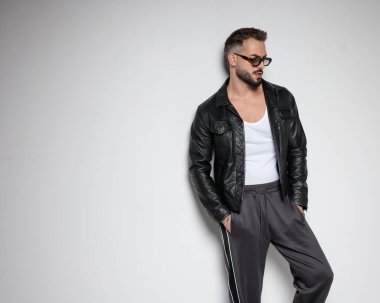 Siyah deri ceketli kendine güvenen genç adam ceplerinde el ele tutuşuyor, başka tarafa bakıyor ve gri arka planda poz veriyor.