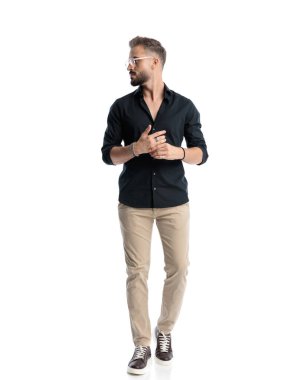 Siyah tişörtlü yakışıklı bir adam beyaz arka planda parmaklarını birbirine değdirirken yürüyor ve yan tarafa bakıyor.