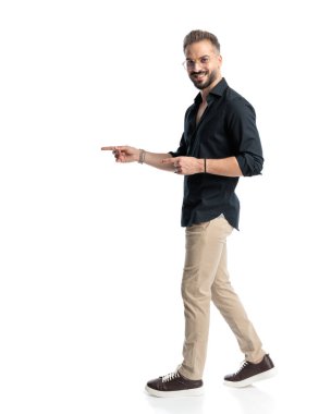 Gözlüklü, parmaklarını sağa sola gösteren ve beyaz arkaplanın önüne adım atan sıradan genç adamın yan görüntüsü