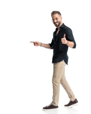 Gözlüklü mutlu genç adam parmağını öne ve yana uzatıyor, gülümsüyor ve beyaz arka planda yürüyor.