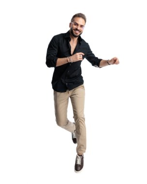 Gözlüklü mutlu genç adam ileriye bakıyor, gülümsüyor ve beyaz arka planın önünde havaya zıplıyor.