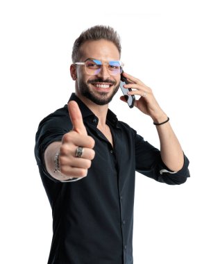 Gözlüklü mutlu genç adam beyaz arka planda baş parmak hareketi yapıyor ve telefonla konuşuyor.