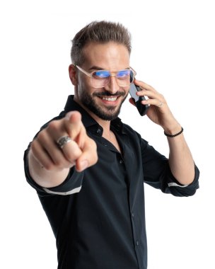 Gözlüklü çekici bir genç telefonda konuşuyor ve beyaz arka planda gülümserken parmağıyla ileri işaret ediyor.