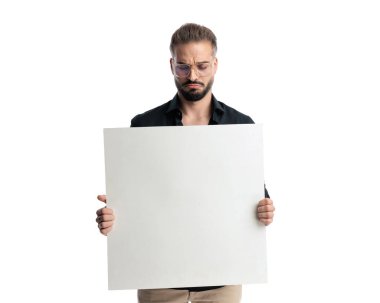 Gözlüklü sıradan bir genç adam tahtayı tutarken ve beyaz arka planda dururken suratını asıyor.