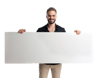 Gözlüklü mutlu genç adam boş beyaz tahtayı tutuyor ve beyaz arka planda sunum yaparken gülümsüyor.