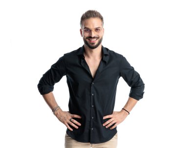 Gözlüklü, kalçasında el ele tutuşan ve beyaz arka planda ileriye bakarken gülümseyen mutlu genç bir adam.