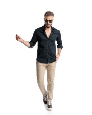 Güneş gözlüklü seksi genç adam parmaklarını şaklatıyor ve beyaz arka planda yürüyor.