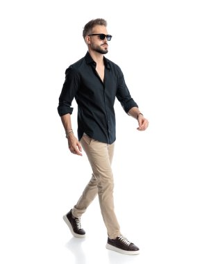 Güneş gözlüklü seksi genç adam kendine güvenerek yürüyor ve beyaz arka planda başka tarafa bakıyor.