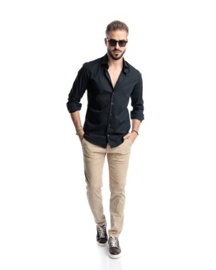 Güneş gözlüklü havalı sakallı adamın uzun bir resmi. Beyaz arka planda elleri ceplerinde ileri doğru hareket ediyor.