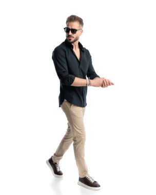 Güneş gözlüklü, beyaz arka planda yürüyen ve avuçlarını ovuşturan havalı genç adamın yan görüntüsü